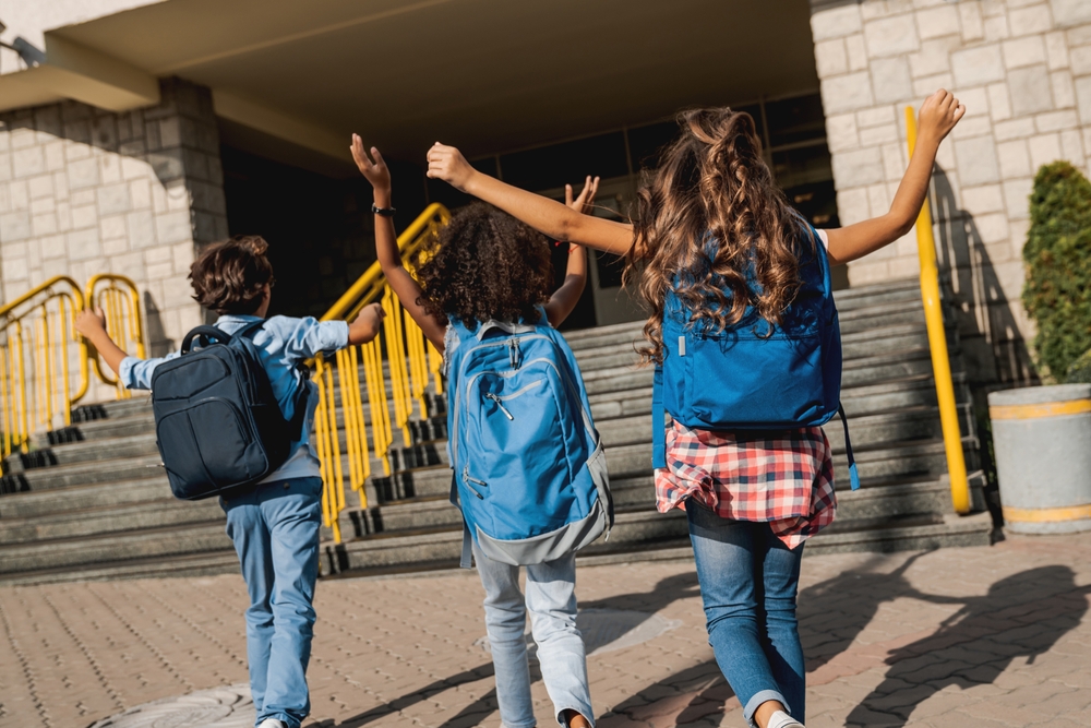 Three school-aged children wearing properly fitted backpacks walk toward a school building, promoting backpack safety and spinal health for kids.